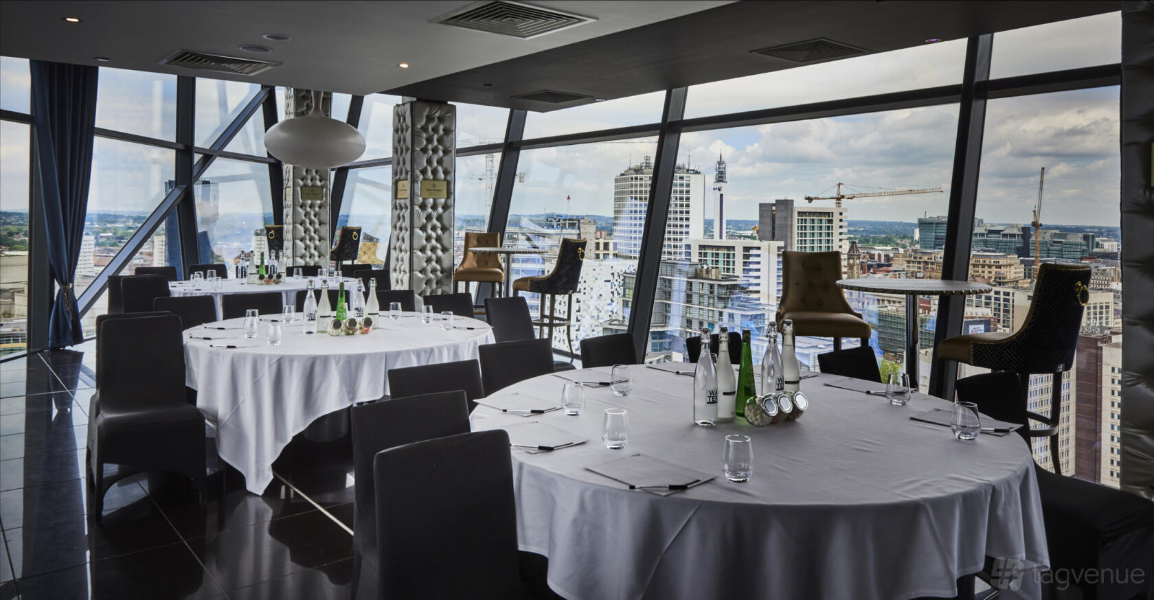 A function room with floor-to-ceiling windows, round tables with white linens, and city skyline views at The Cube.