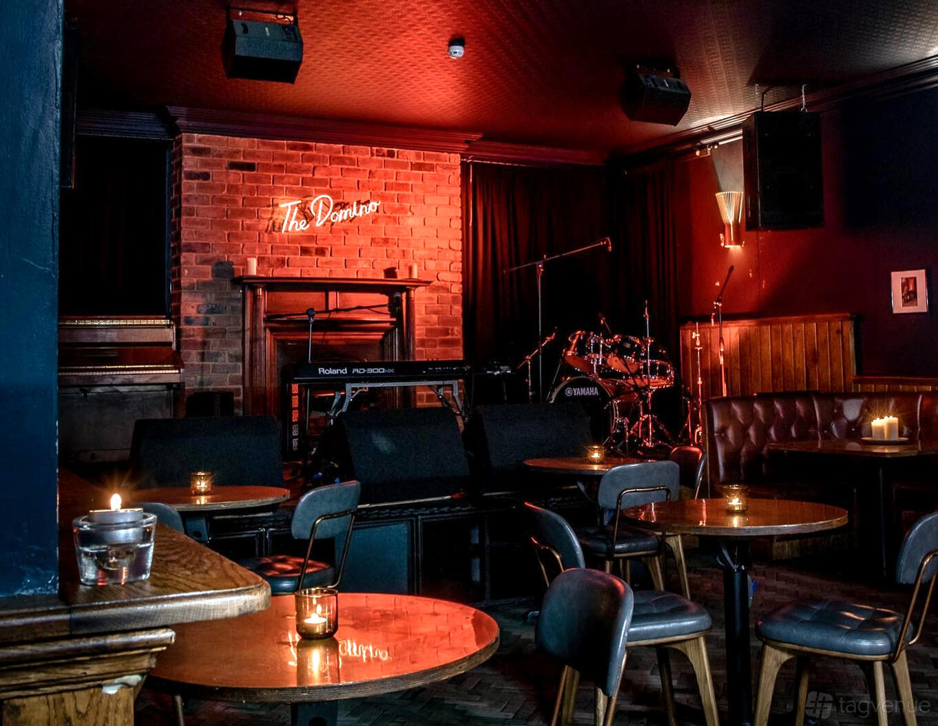 An event space in a cafe with exposed brick walls, a small stage, musical instruments, and round candlelit tables at The Domino Club.
