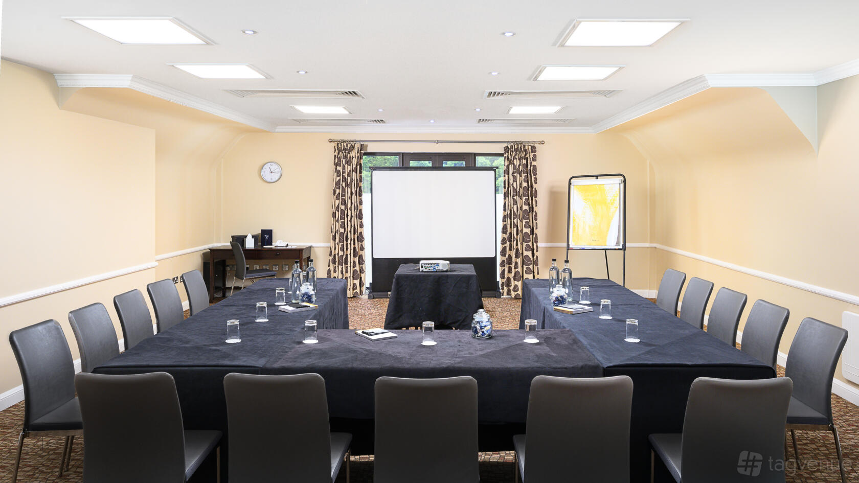 A meeting room with U-shaped black tablecloths, grey chairs, and a projector screen at Voco Oxford Spires.