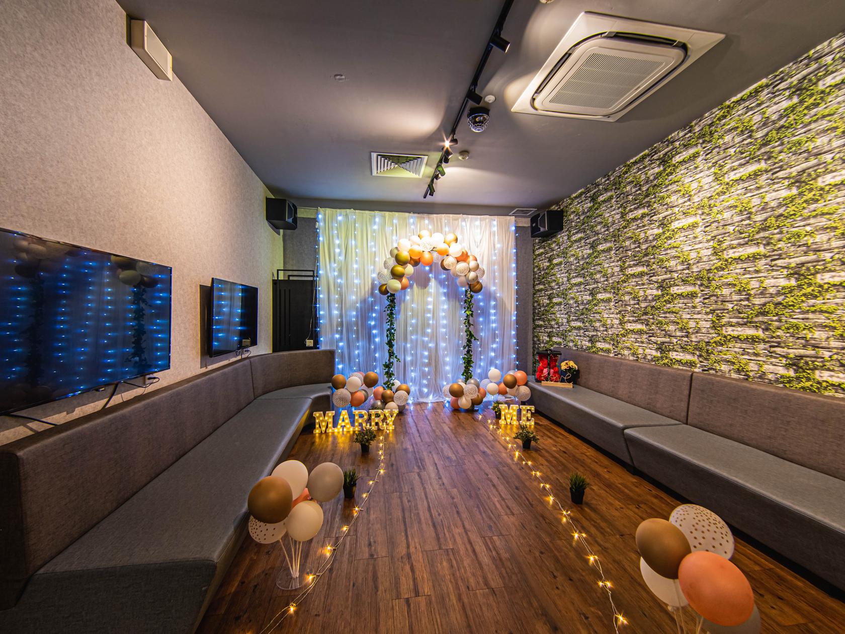 A function room with long grey sofas, a balloon arch with fairy lights, and a TV at 7th Heaven KTV Cafe.