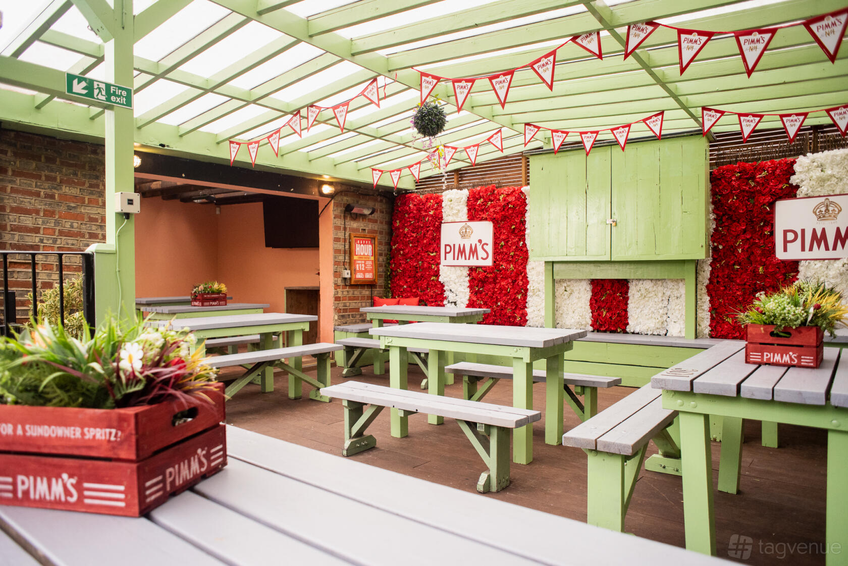 A covered terrace at a pub with pastel green picnic benches, floral walls, and decorative bunting at The Abbey Tavern.