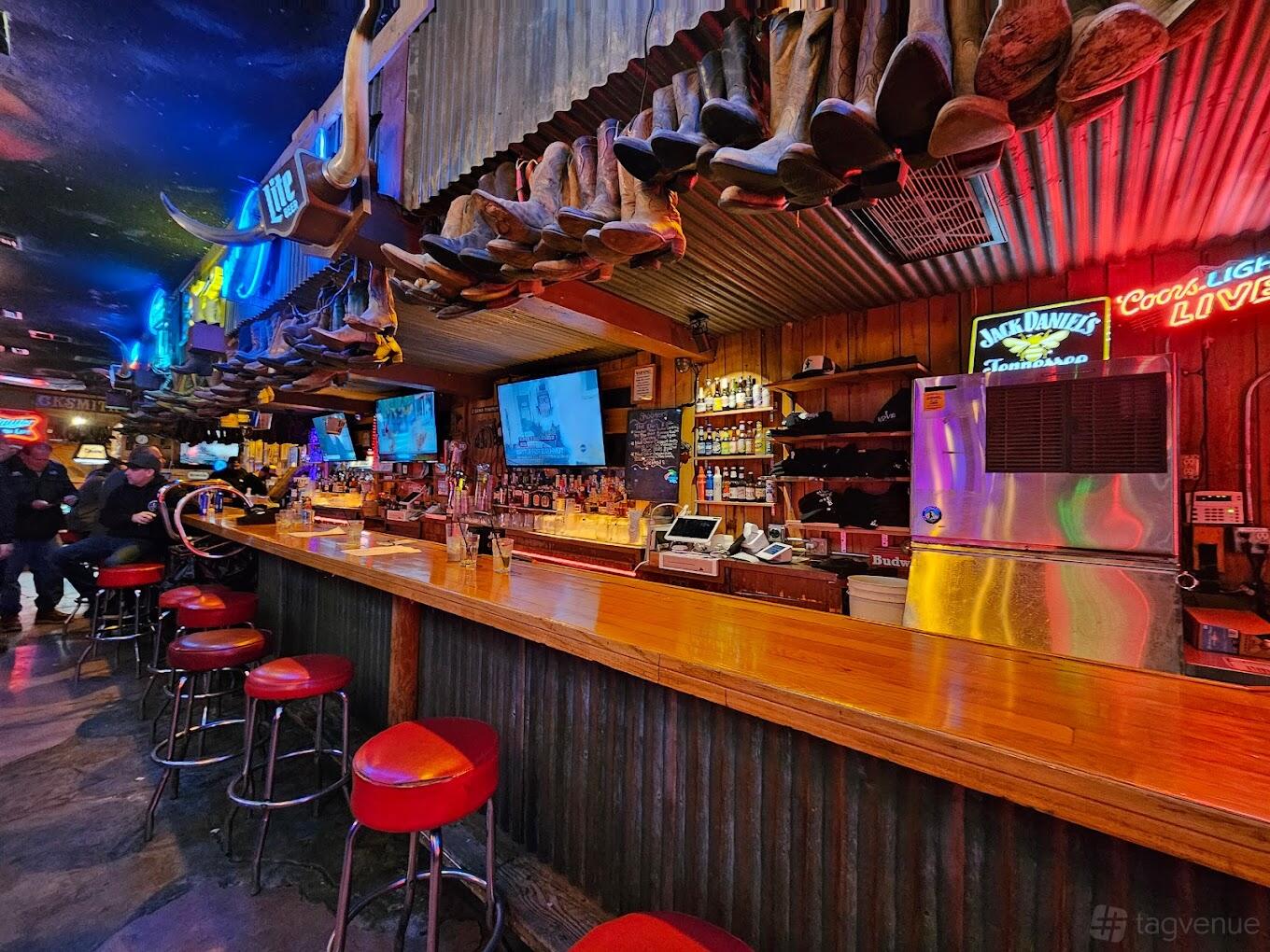 A bar with a long wooden counter, red stools, neon signs, and cowboy boots lining the ceiling at Cowboy Palace Saloon.