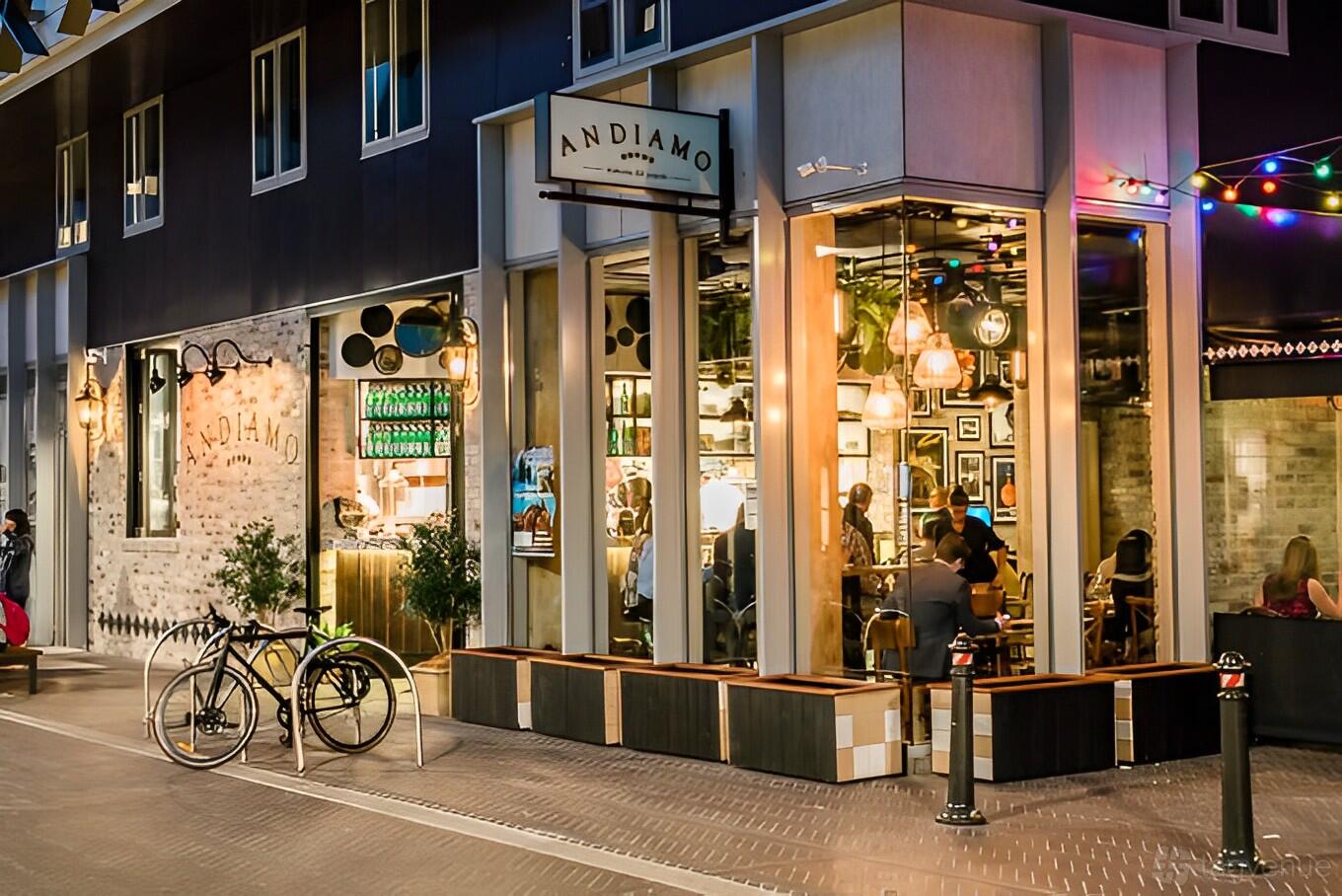A restaurant exterior with large glass windows, warm lighting, visible diners, and outdoor benches at Andiamo Trattoria Chippendale.