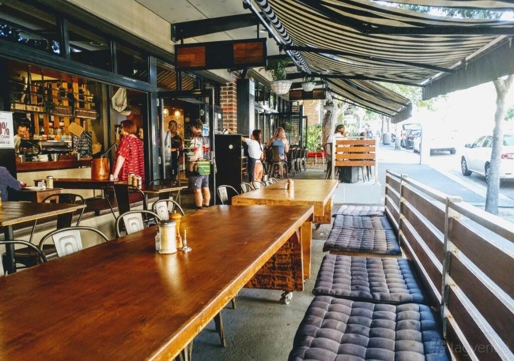 A terrace at a cafe with wooden tables, cushioned bench seating, and striped retractable awnings at Hoochie Mamma.