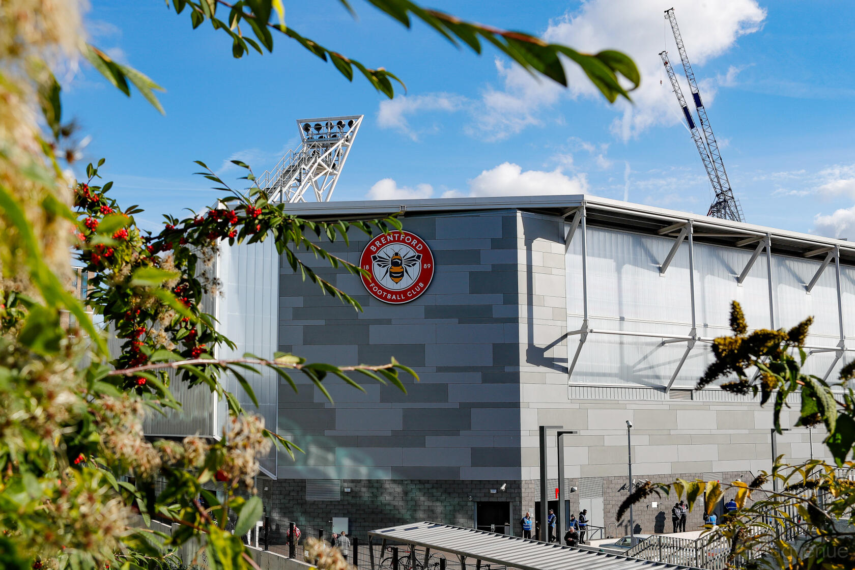 A stadium exterior with Brentford Football Club signage, metal panel walls, and stadium lighting towers.