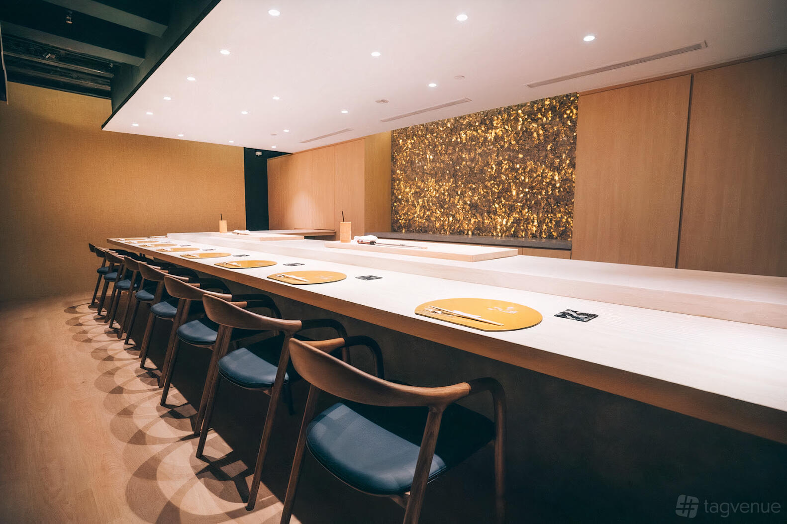 A restaurant with a long wooden sushi counter, modern chairs, and a golden textured feature wall at Ikkoten Omakase.