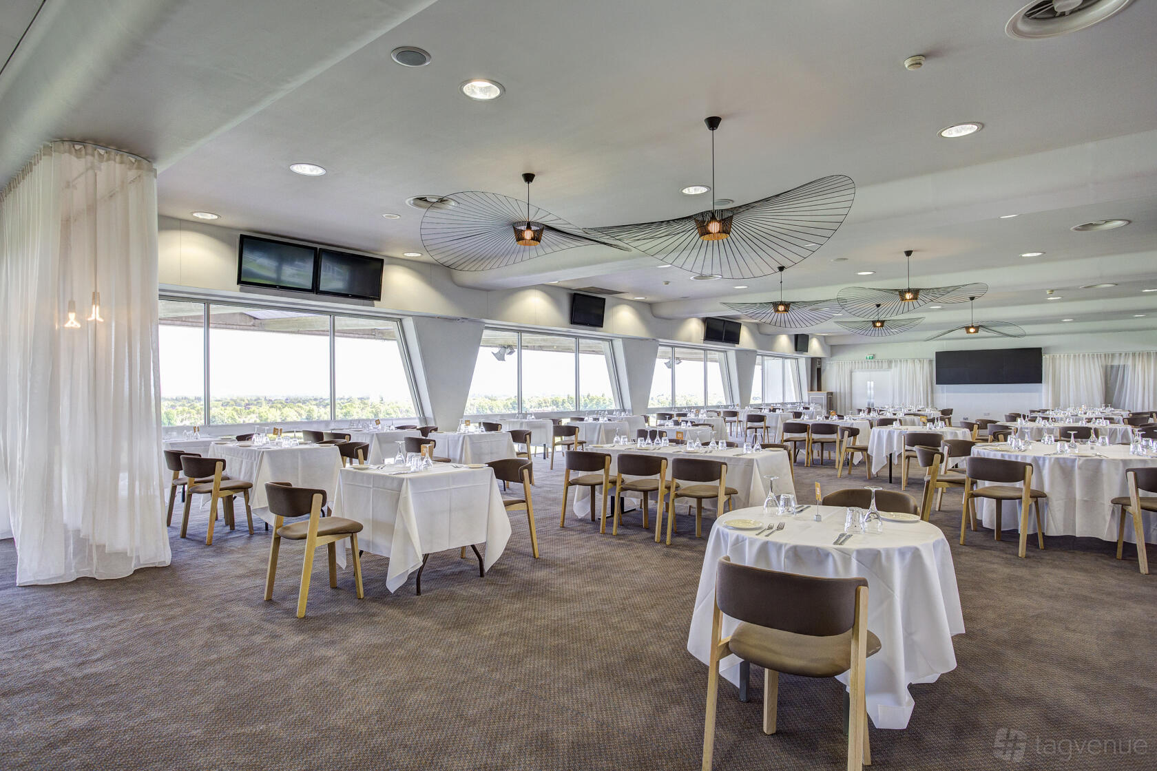 An event venue with floor-to-ceiling windows, round tables with white linens, and sculptural ceiling lights at Sandown Park Racecourse.