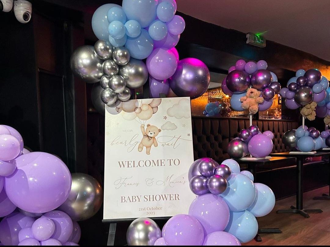 A bar with tufted leather banquette seating, metallic and pastel balloon decorations, and a baby shower welcome sign at Basement 144.