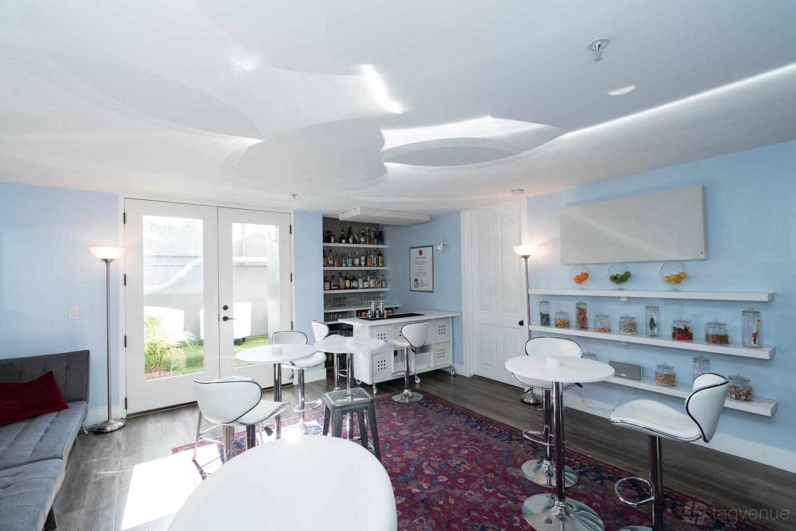 An event venue with white bar tables, acrylic chairs, a stocked bar, and glass jars on shelves at Versatile Art Deco Suite.