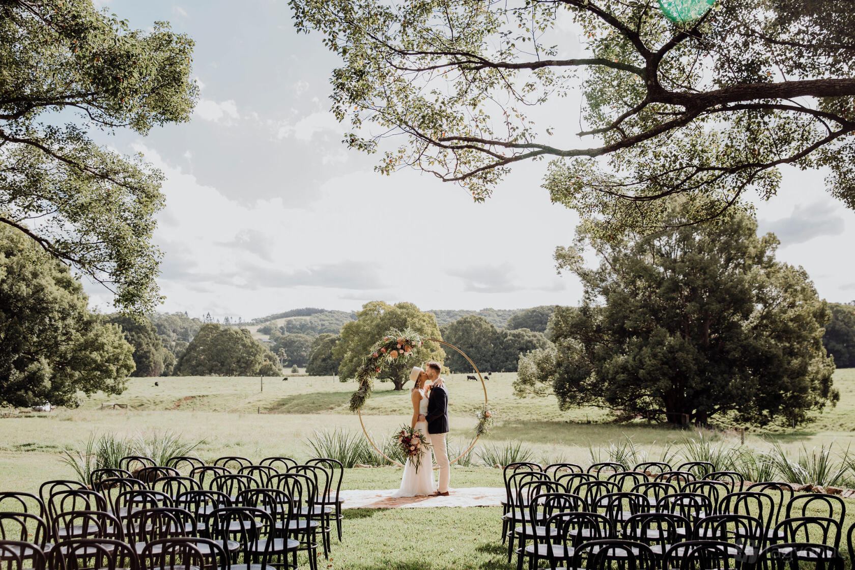 A terrace with rows of black chairs facing a floral arch under large trees at Frida's Field.