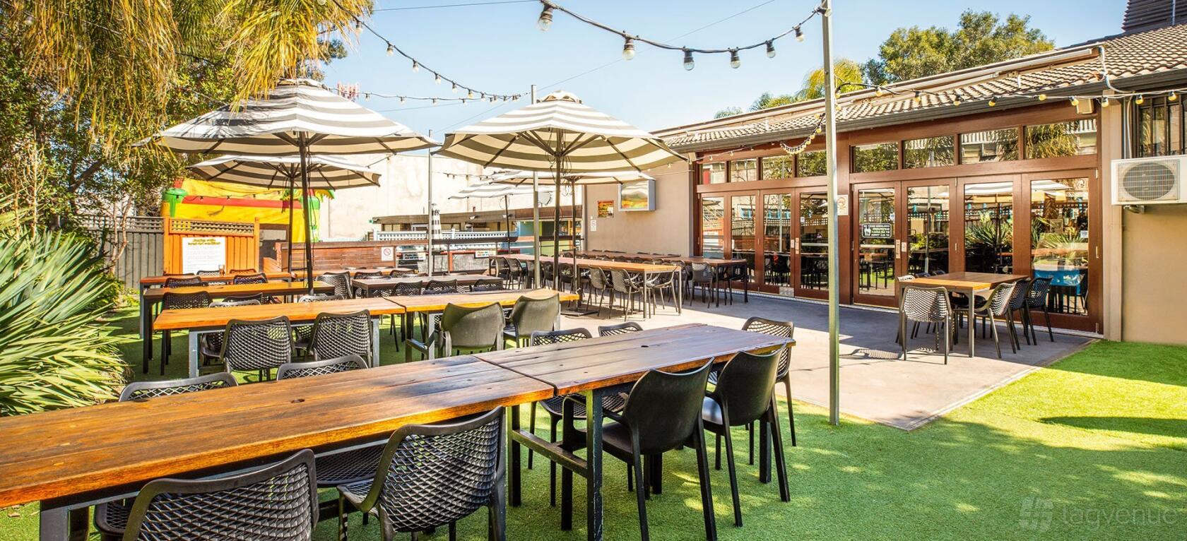 An outdoor hotel patio with wooden tables, black chairs, umbrella shades, and string lights at The High Flyer.
