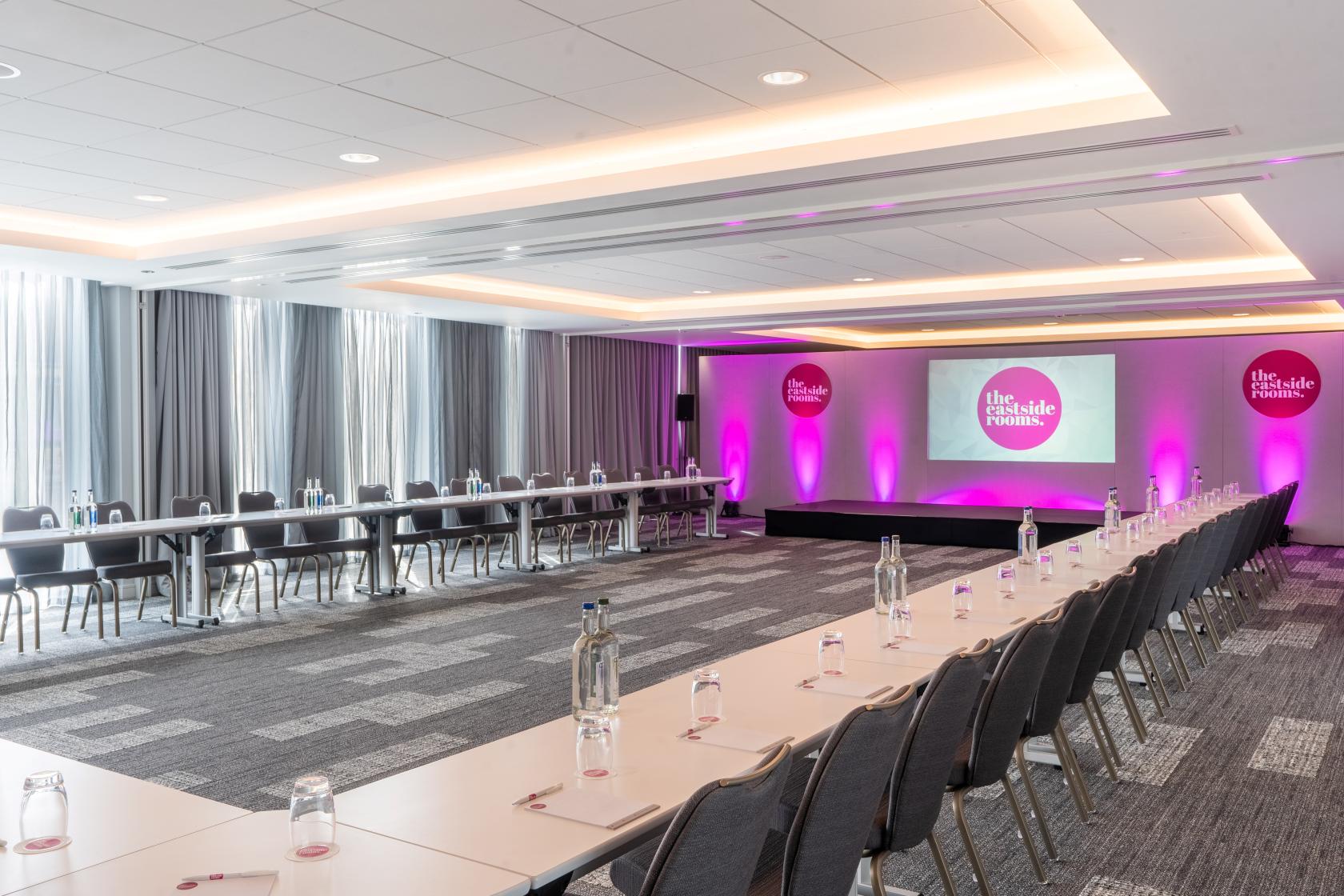 A meeting room with U-shaped tables, bottled water, a projector screen, and purple uplighting at The Eastside Rooms.