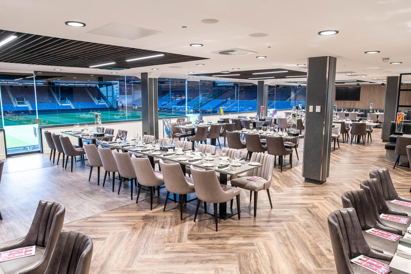 An event suite with banquet tables, upholstered chairs, and pitch views through floor-to-ceiling windows at Burnley Football Club.