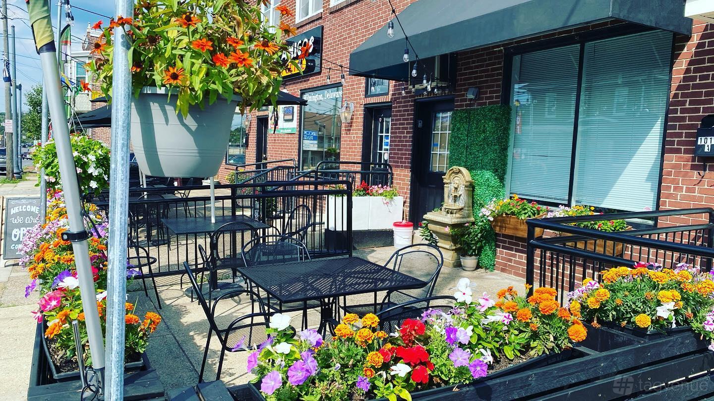 A restaurant terrace with black metal tables, flower planters, and a decorative water fountain at THREE AMIGOS.