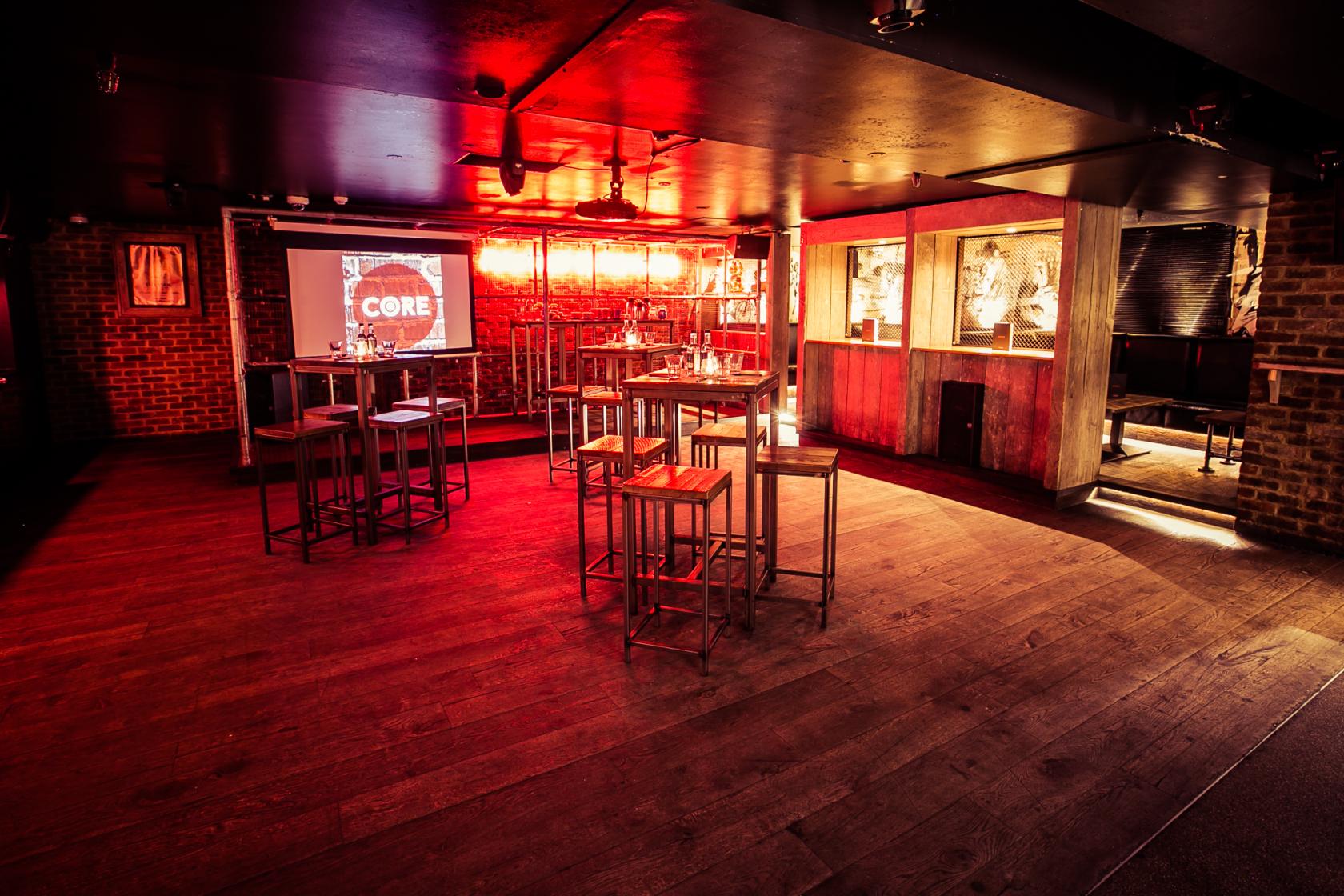 An event space in a bar with wooden floors, high tables, glowing red lighting, and a projection screen at Core.