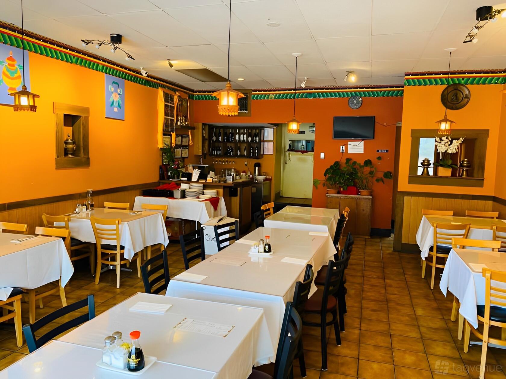 A dining room with white tablecloths, wooden chairs, and orange accent walls at Tashi Delek Party Palace.