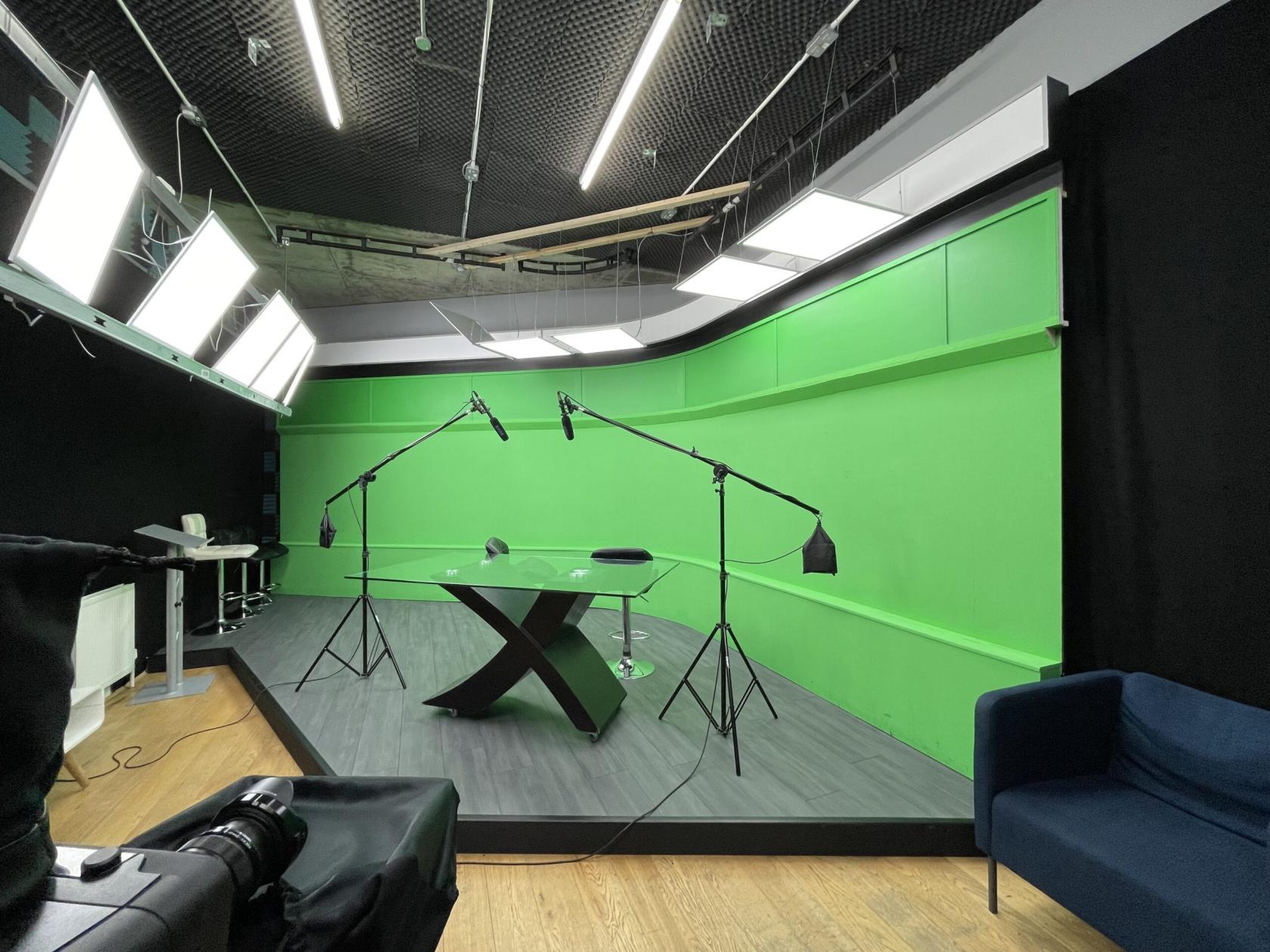 A recording studio with a green screen backdrop, suspended microphones, and a glass table at TMH Studios.