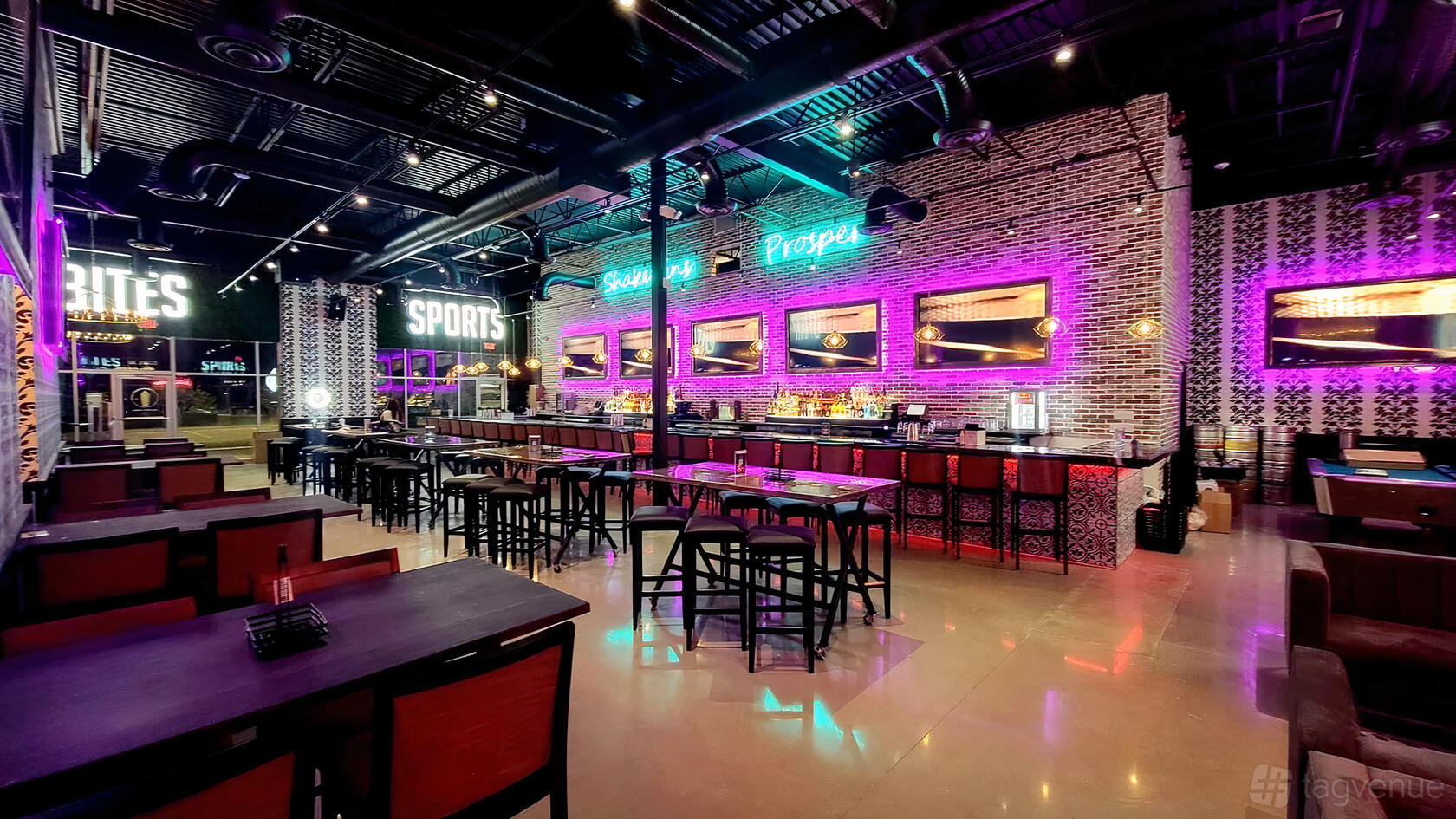 A bar with exposed brick walls, neon signage, high tables, and illuminated shelves at Shakertins - Prosper.
