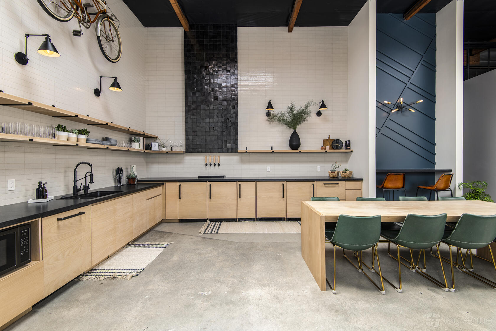 An event space with a long wooden table, green chairs, open shelving, and a kitchenette at TrueWork Environment.