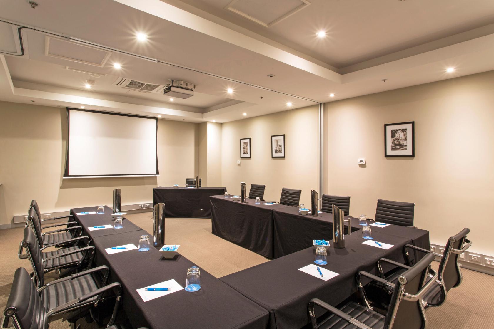 An event space with U-shaped conference tables, black chairs, and a projection screen at Mantra Melbourne Airport.