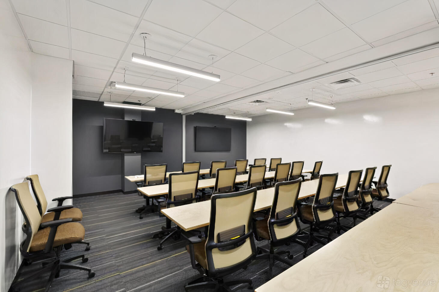 A training room with rows of ergonomic chairs, wood tables, and dual wall-mounted screens at Venture X Dallas by the Galleria.