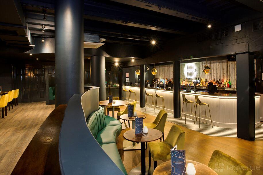 A lounge in a pub with curved green banquettes, small round tables, and a bar with pendant lighting at Slug and Lettuce Liverpool.