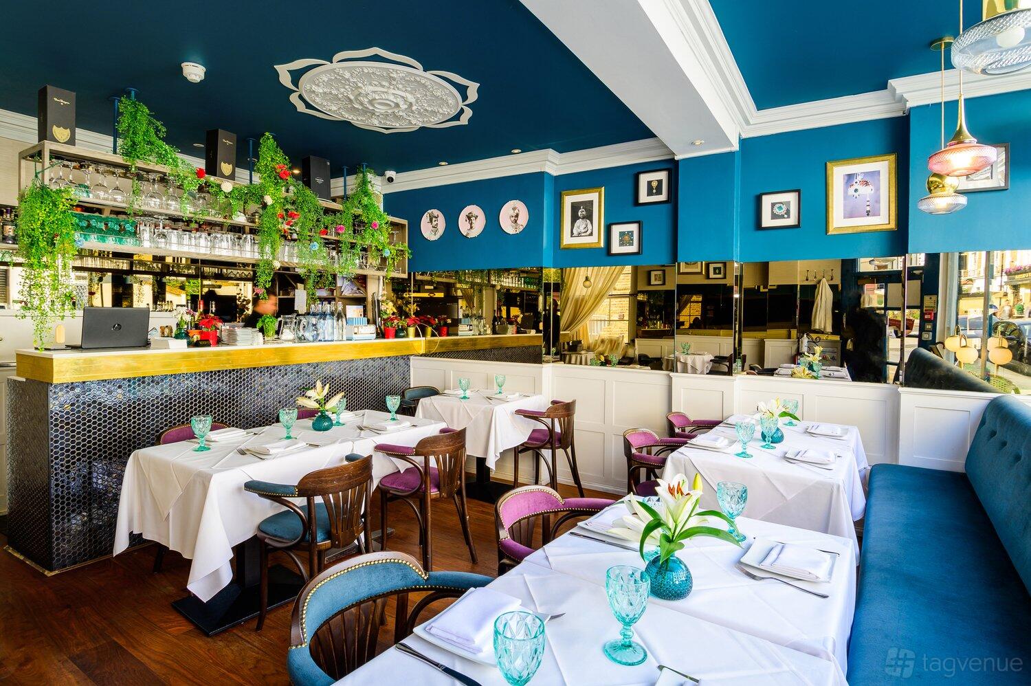 A restaurant with white tablecloths, blue walls, gold-trimmed bar, and hanging greenery at Chakra Kensington.