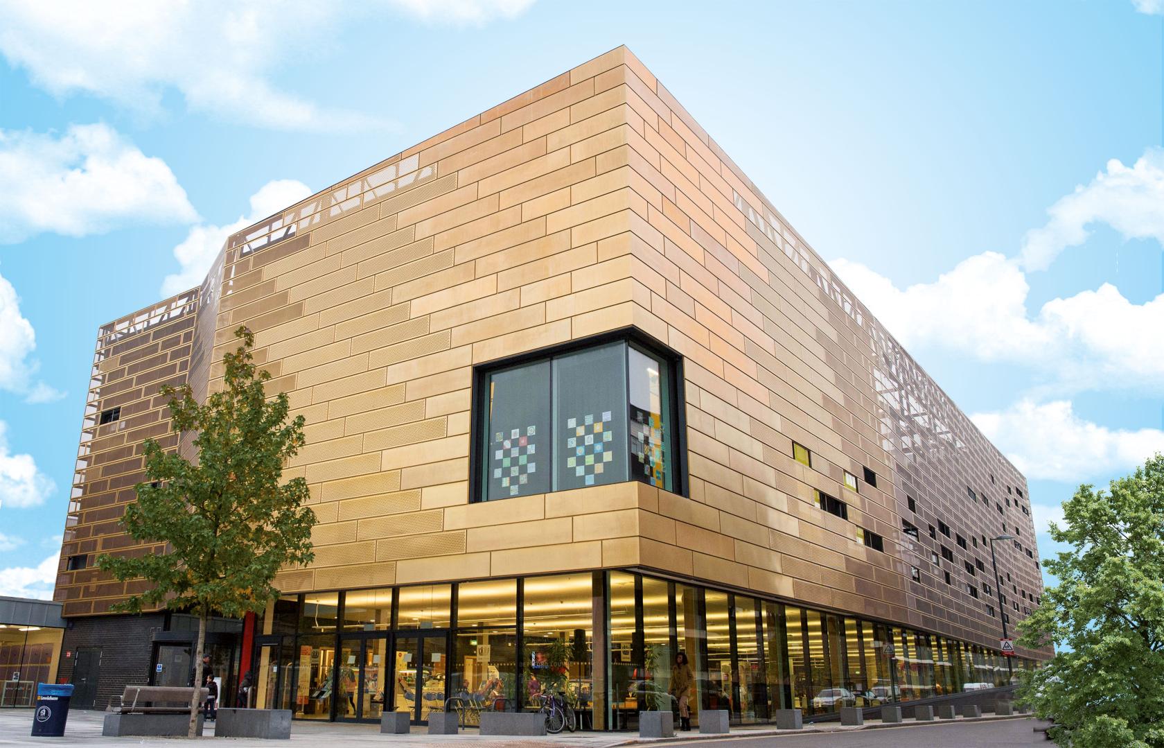 A sports hall with a gold-clad facade, large windows, and trees at Deptford Lounge.