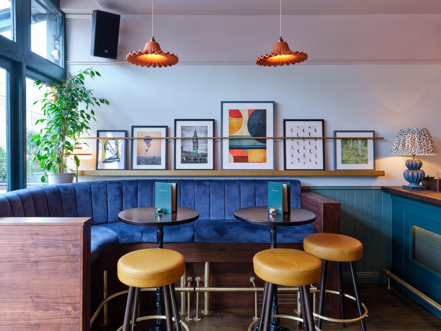 An event space in a bar with a blue velvet booth, round tables, leather stools, and framed artwork at LOCKES Bar Clapham Junction.