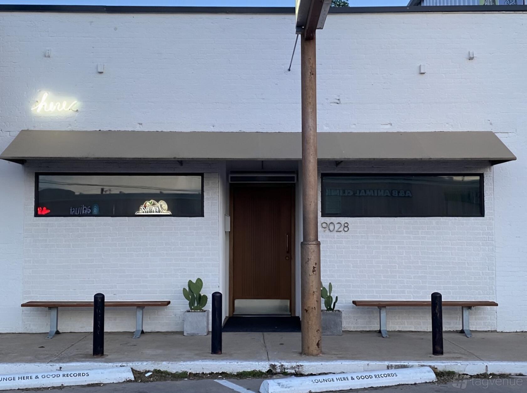 A bar entrance with white brick facade, large front windows, brown door, and benches at Lounge Here.