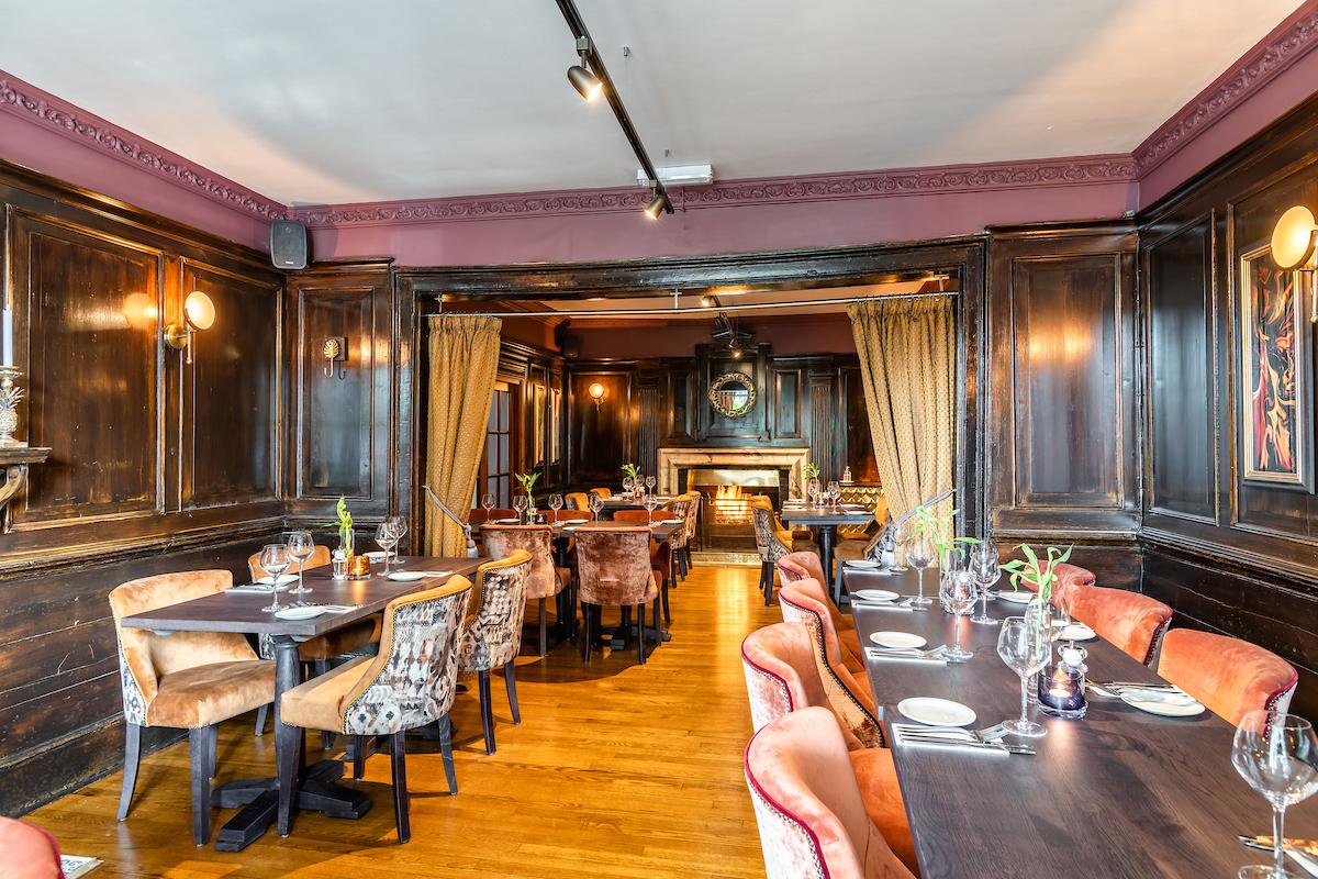 A dining room with dark wood panelled walls, velvet chairs, and set tables at The Bell, Ashton Clinton.