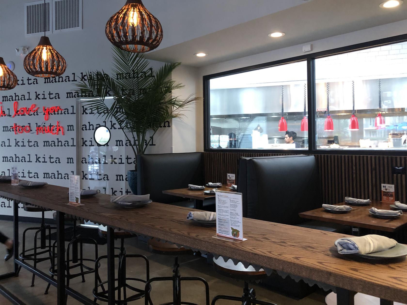A restaurant dining area with wooden tables, black booth seating, pendant lights, and an open view into the kitchen at Be More Pacific.