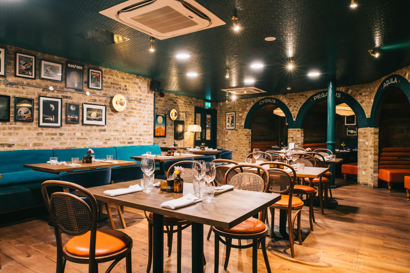 A pub function room with exposed brick walls, framed artwork, wooden tables, and blue banquette seating at The Astronomer.
