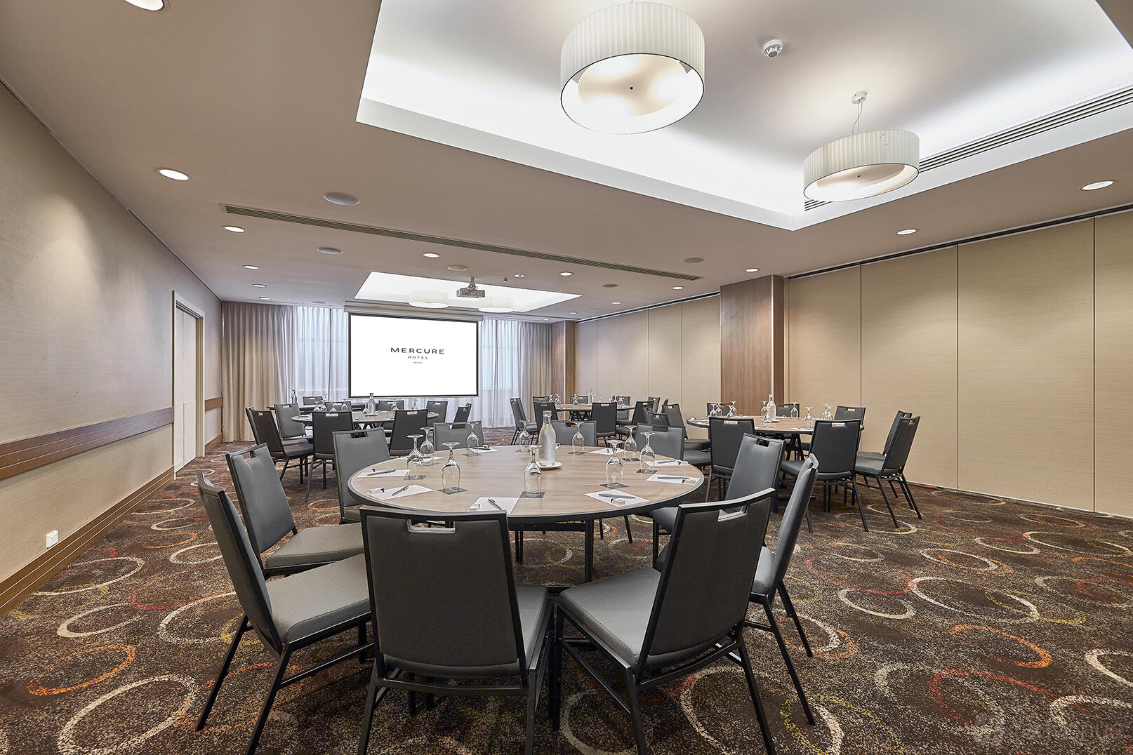 A meeting room with round tables set with water glasses, ceiling lights, and a projector screen at Mercure Perth.