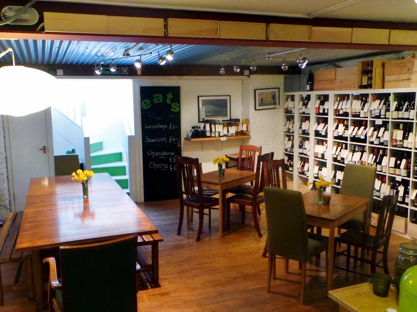 A bar with wooden tables, mixed chairs, and shelves filled with wine bottles at D Vine Cellars.