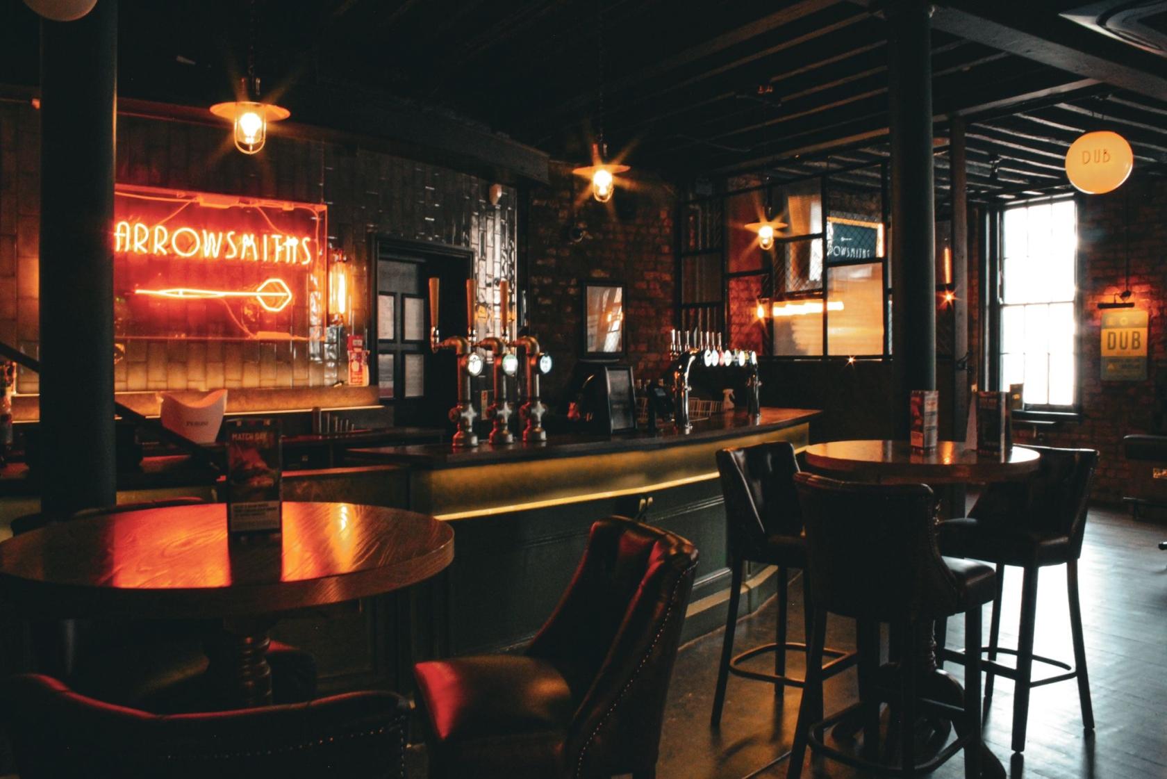 A pub with exposed brick walls, neon signage, round wooden tables, and leather chairs at Arrowsmiths @ The Shipping Forecast Liverpool.