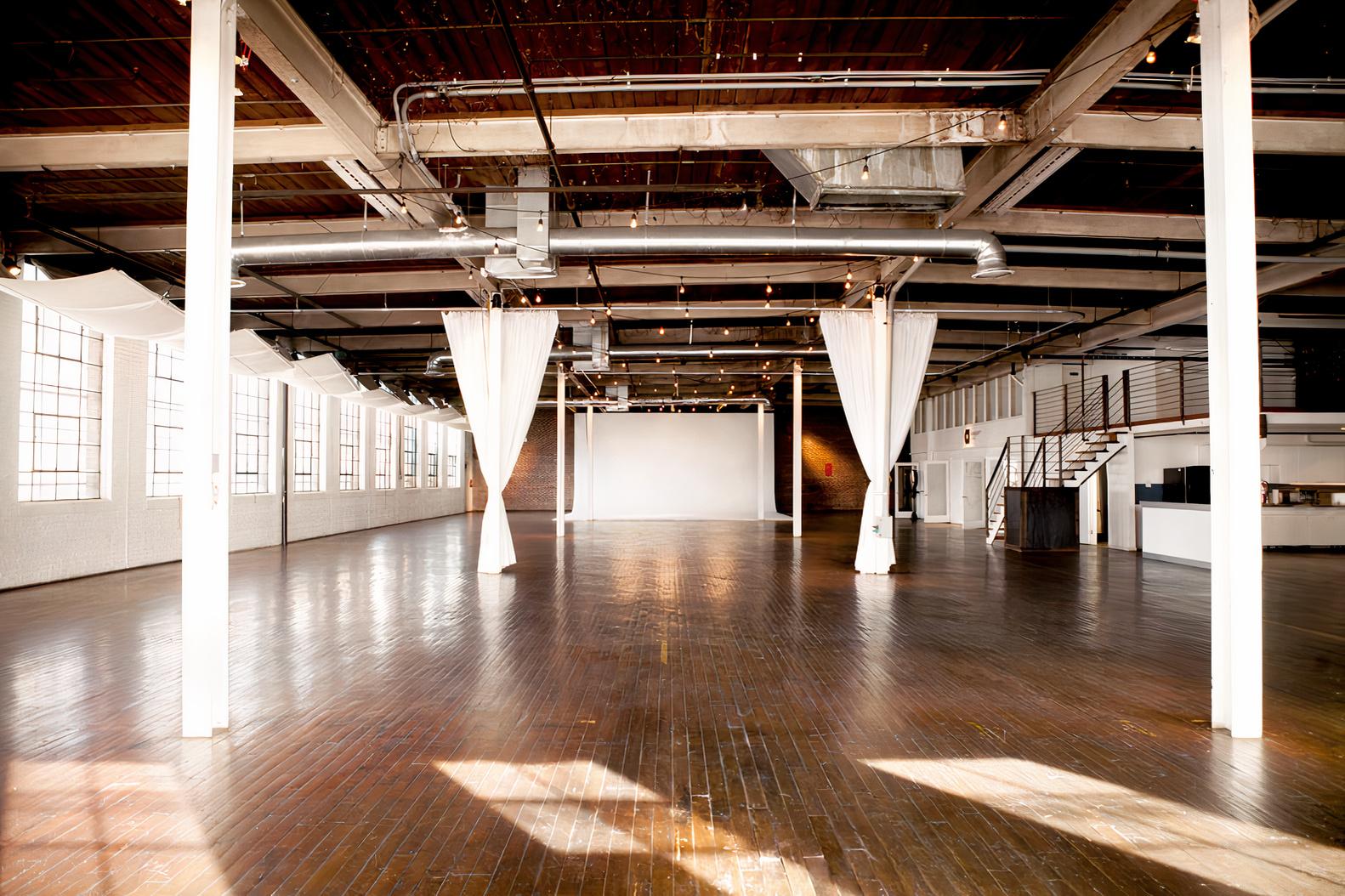 An event space with large windows, polished wooden floors, white draped columns, and exposed beams at Ambient + Studio.