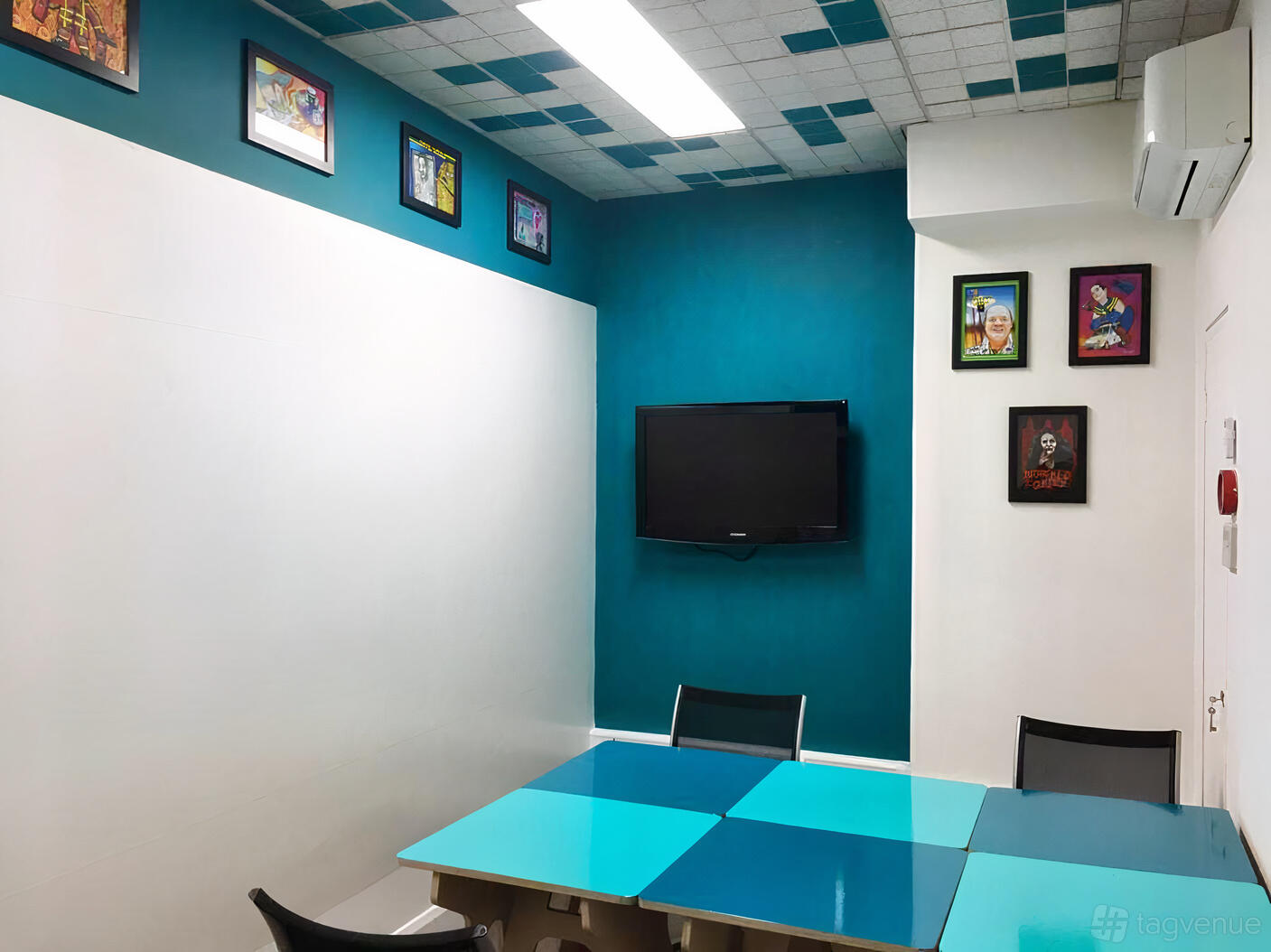A meeting room with teal accent walls, colorful framed artwork, and a wall-mounted TV at tcube.
