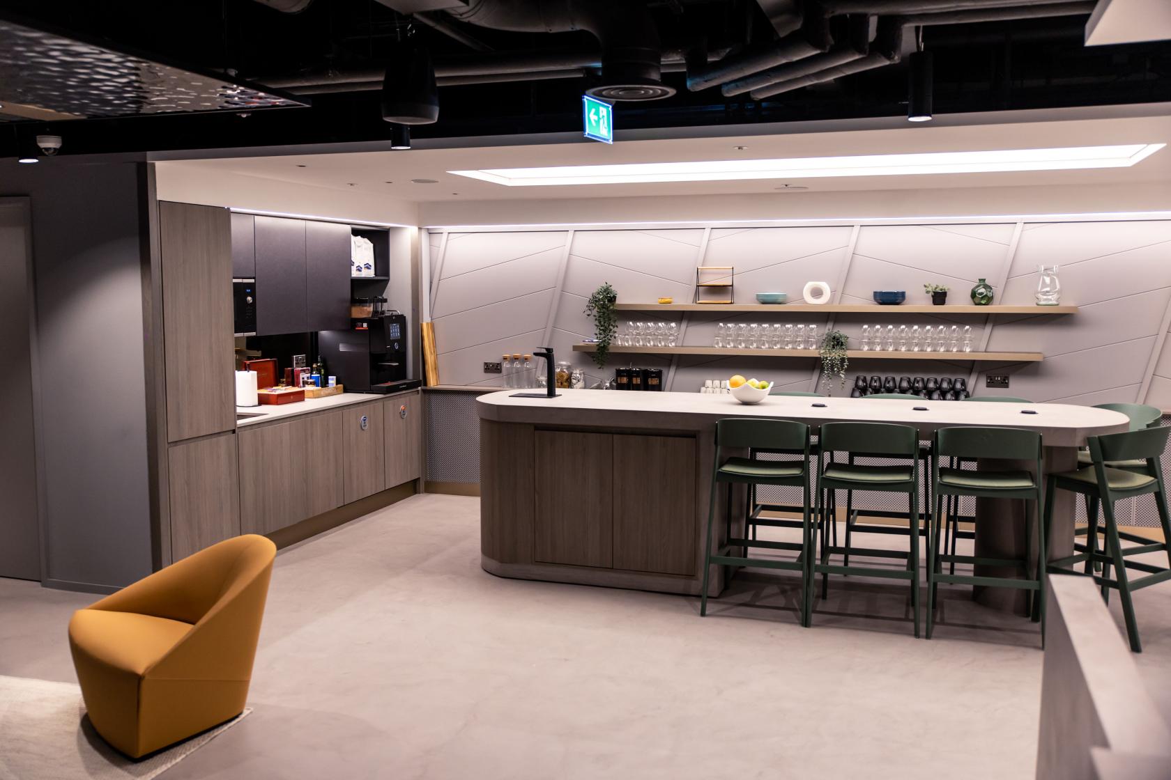 An event venue with a long bar counter, open shelving, green barstools, and a kitchenette at UNTIL Liverpool Street.