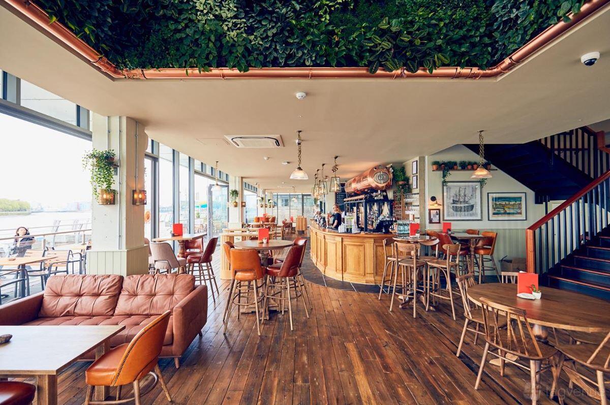 A pub bar with wood flooring, leather chairs, high tables, and a ceiling decorated with hanging greenery at The Oystercatcher.