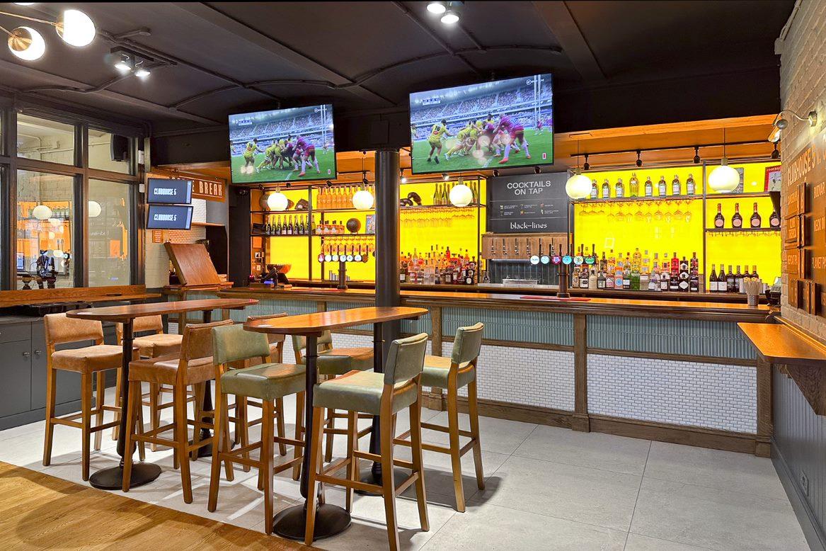 An event space in a bar with high tables, wooden stools, illuminated shelves, and two TVs at The Three Lanterns.
