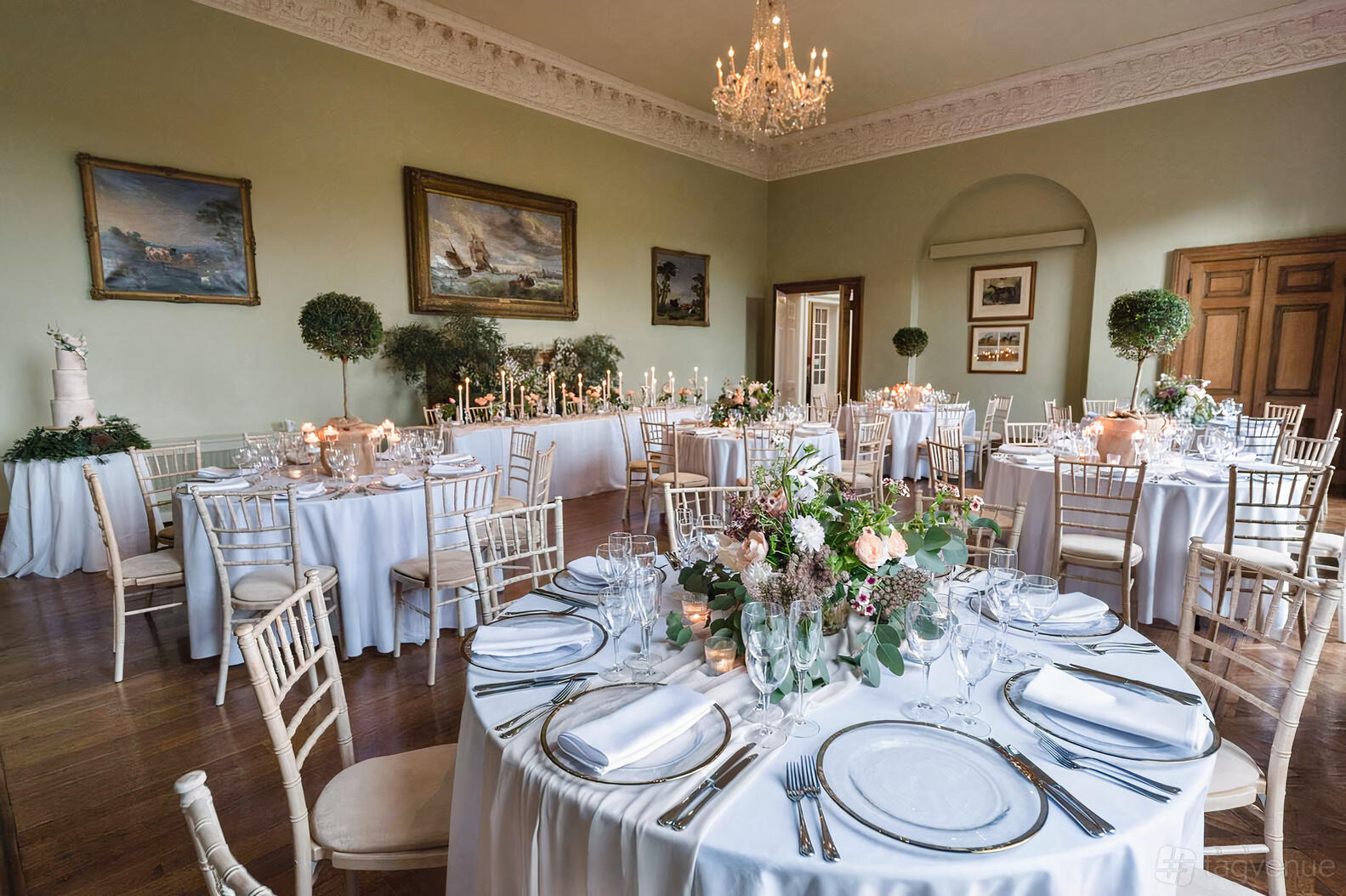 A hall with round tables draped in white linens, floral centerpieces, chandeliers, and framed artwork at Kelmarsh Hall.