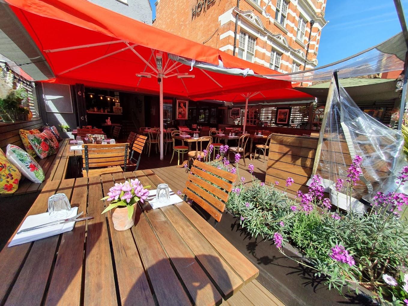 An outdoor restaurant patio with wooden tables, vibrant red umbrellas, floral cushions, and potted flowers at Bacco Ristorante Italiano.