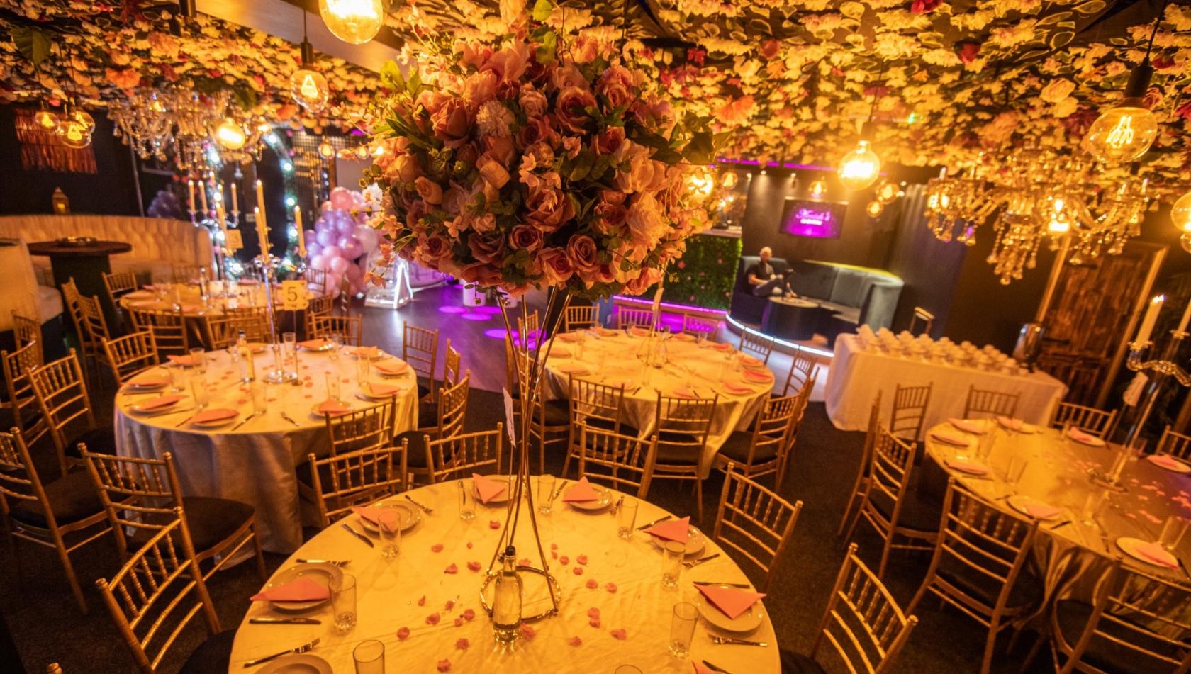 An event space with floral ceiling decor, gold chairs, round tables with pink napkins, and chandeliers at Memoirs Banqueting.