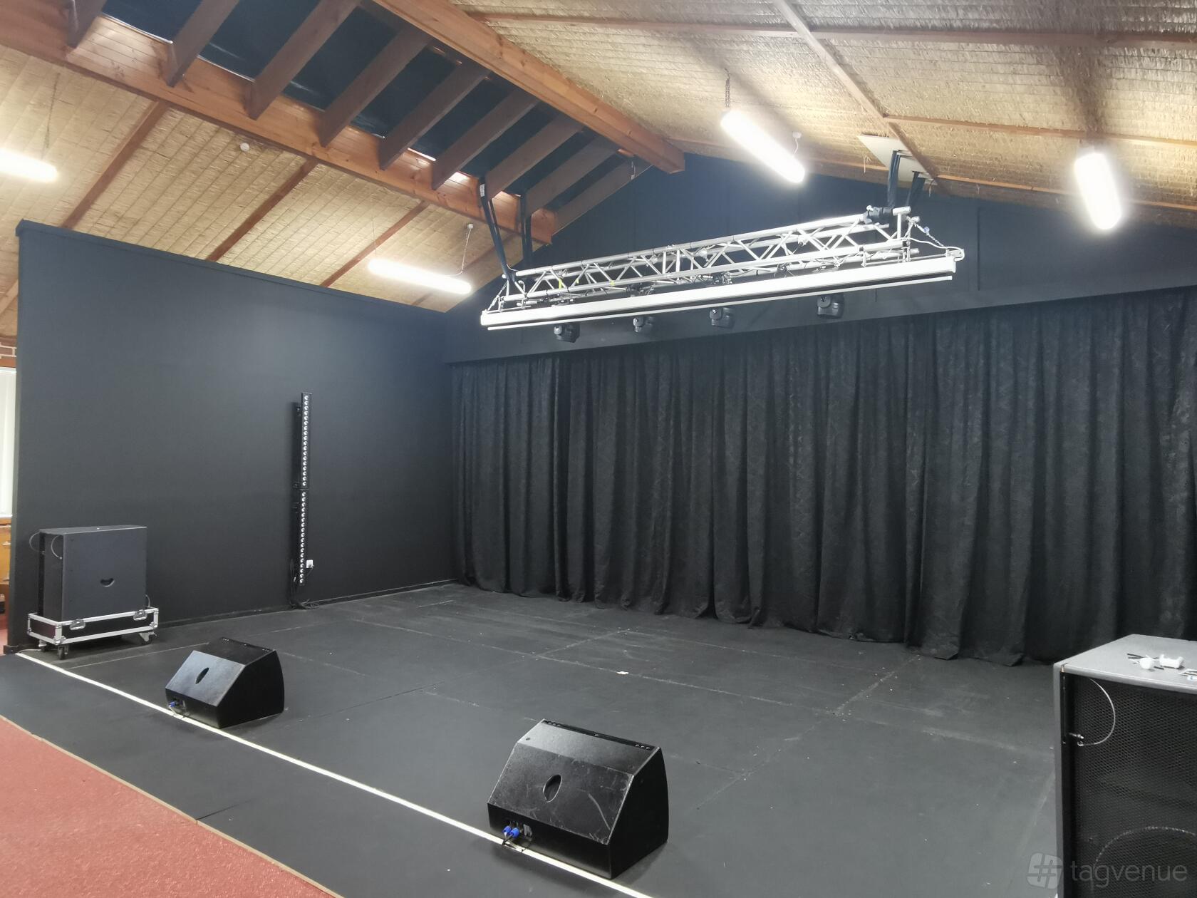 An auditorium stage with black curtains, overhead lighting rig, and speakers at Sinofield Edu Retreat.