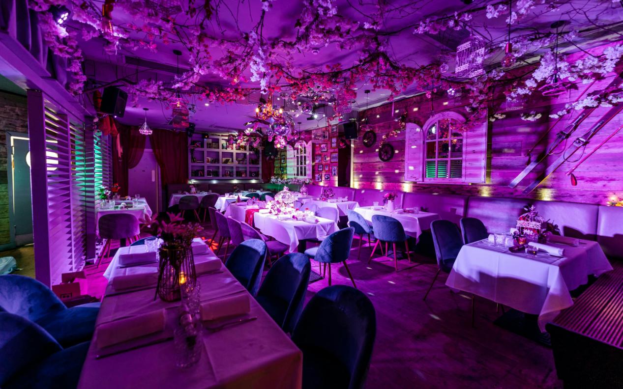 A bar with floral ceiling decor, purple lighting, and white tablecloth dining tables at The Chelsea Lodge.