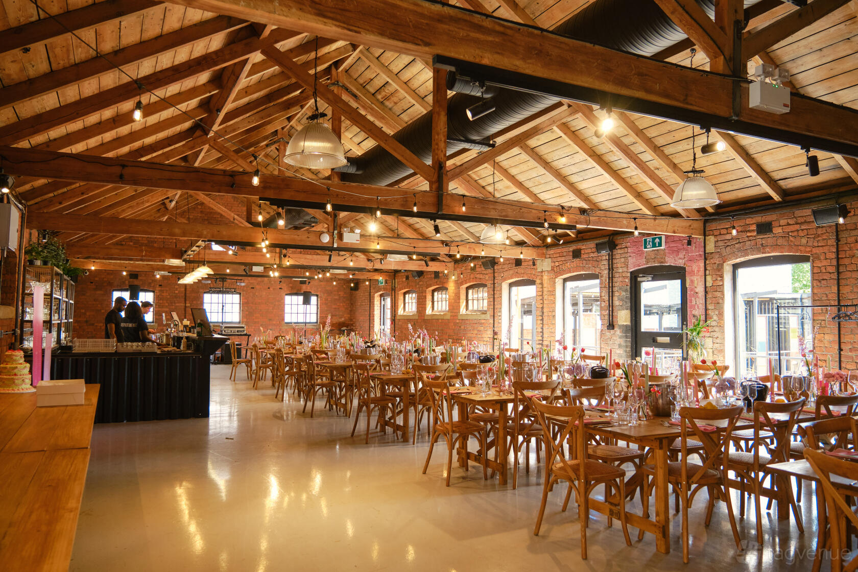 An event venue with exposed brick walls, wooden beams, and banquet tables with wooden chairs at The Stables.