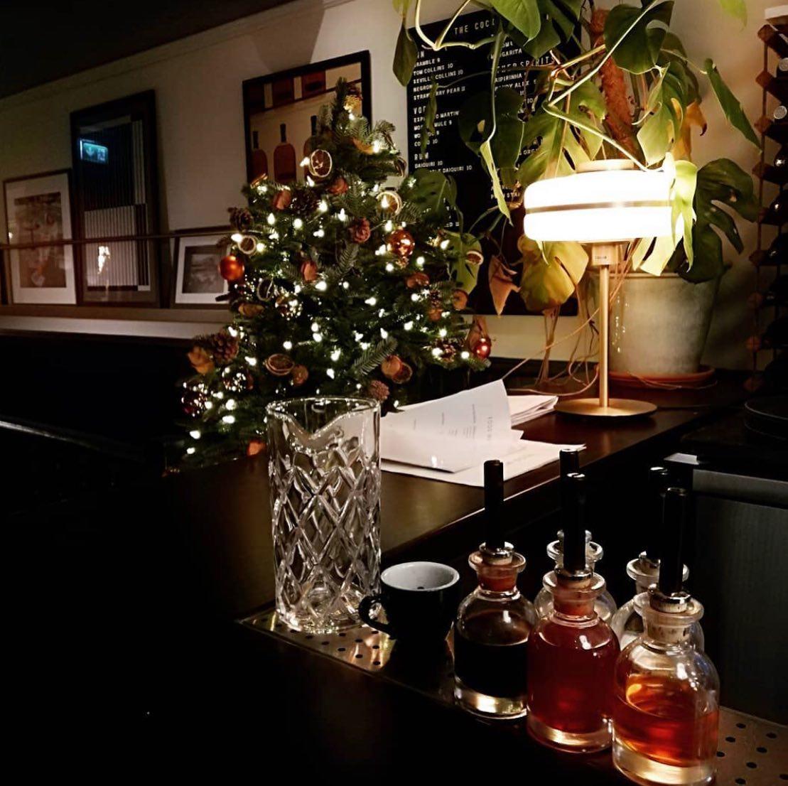 An event space in a cocktail bar with a decorated Christmas tree, glass mixing pitcher, and illuminated table lamp at LOCKES Bar Covent Garden.