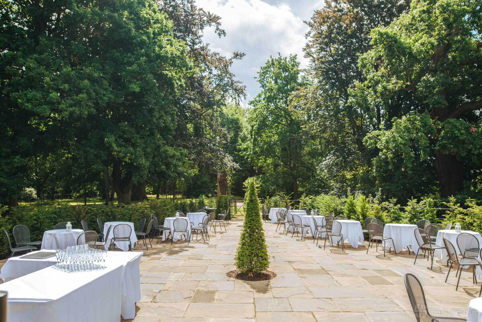An outdoor event venue with stone patio, scattered tables draped in white linens, and surrounding greenery at Pembroke Lodge.