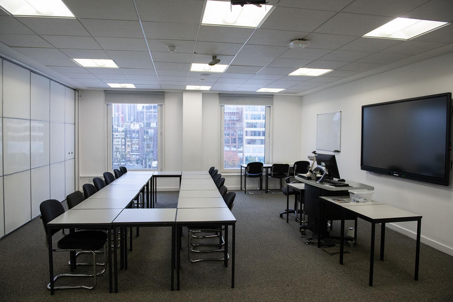 A meeting room with large windows, U-shaped tables, office chairs, and a wall-mounted screen at University of Law - London Moorgate.